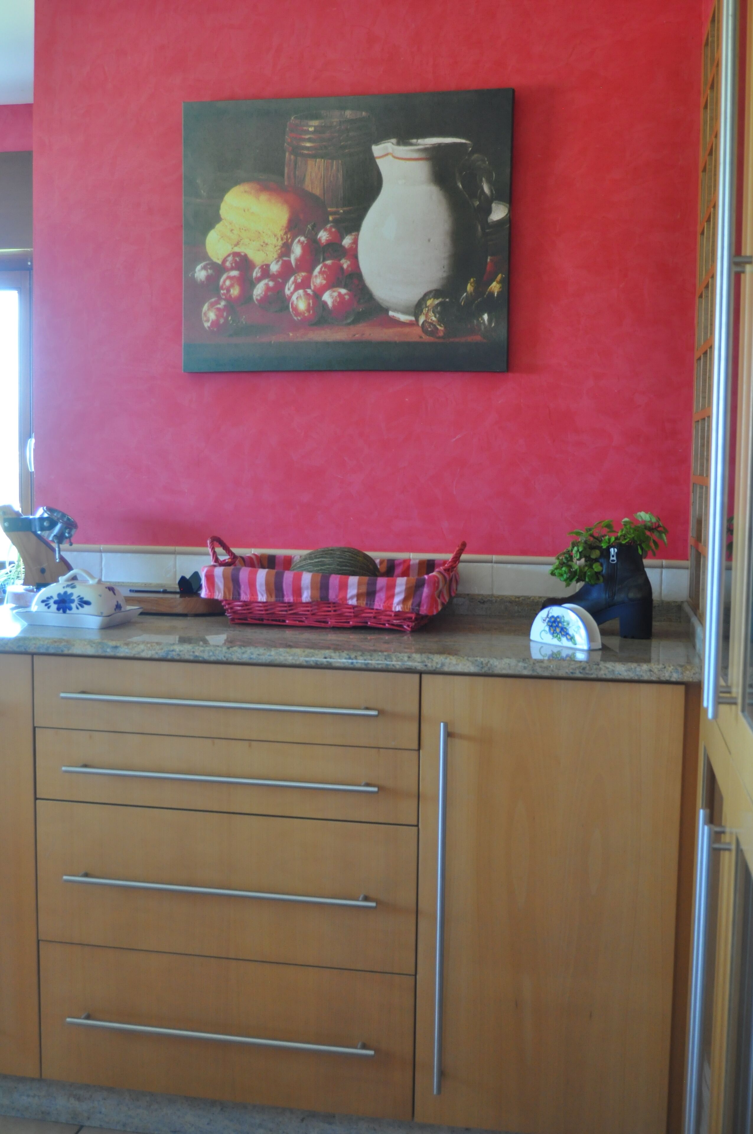 A kitchen with a red wall, a painting of fruits and a jug, a wicker basket, and a potted plant.