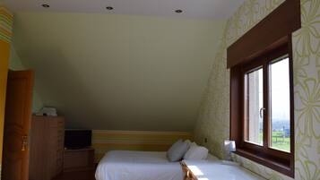 A bedroom with two beds, a wooden wardrobe, a television on a stand, and a window with a view of greenery.