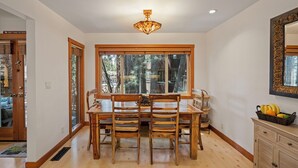 A dining area with a wooden table and chairs, a chandelier, a large window with a view of trees, and a mirror on the wall.