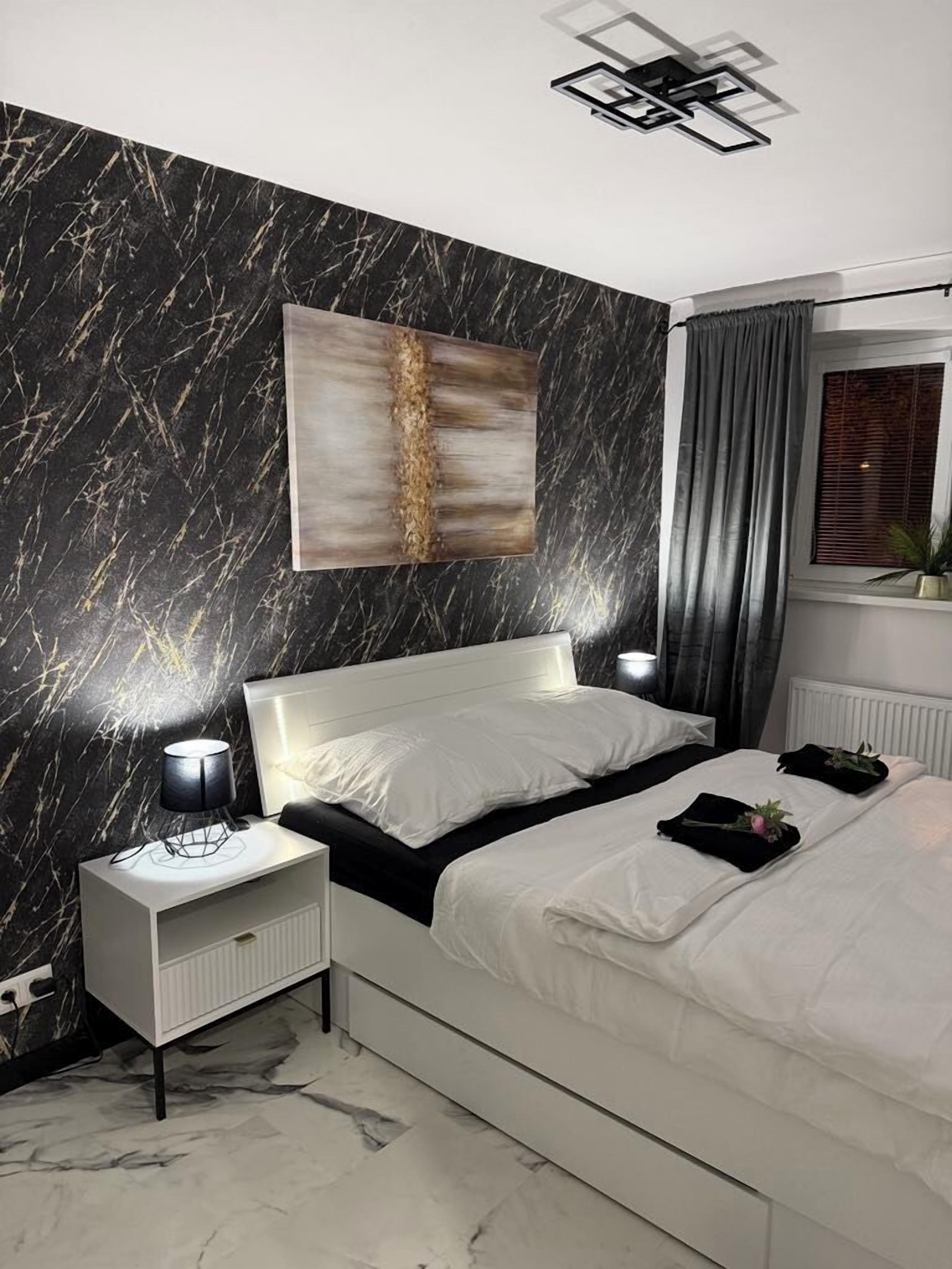 Une chambre moderne avec un mur à motifs de marbre, un lit avec une literie blanche, une table de chevet avec une lampe et une fenêtre avec des rideaux.
