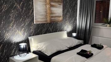 Een moderne slaapkamer met een wand met marmerprint, een bed met witte beddengoed, een nachtkastje met een lamp en een raam met gordijnen.
