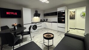 Un salon moderne avec un canapé noir, une table basse ronde et un coin repas comprenant une table et des chaises noires. On aperçoit également une cuisine avec des meubles blancs et un plan de travail noir.