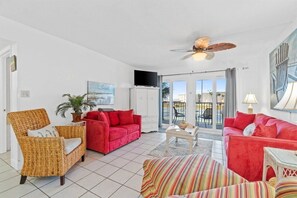 A living room with a red sofa, a wicker chair, a television, and a ceiling fan. - 2BR Condo Sleeps 6 – Pool & Steps to the Beach (Panama City Beach)
