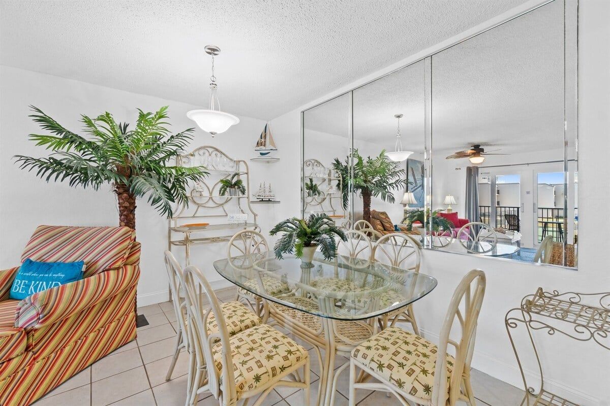 A dining area with a glass table surrounded by wicker chairs, a striped sofa with a blue pillow, and a large mirror reflecting the room.