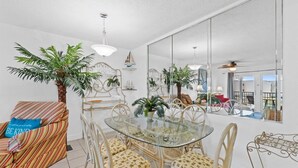 A dining area with a glass table surrounded by wicker chairs, a striped sofa with a blue pillow, and a large mirror reflecting the room. - 2BR Condo Sleeps 6 – Pool & Steps to the Beach (Panama City Beach)