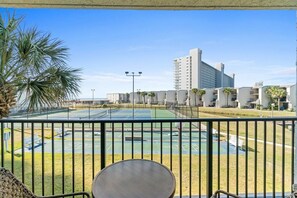 A balcony with a view of a tennis court, palm trees, and a high-rise building in the background. - 2BR Condo Sleeps 6 – Pool & Steps to the Beach (Panama City Beach)