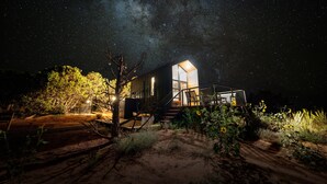 Una casa moderna con un gran ventanal y una terraza de madera, rodeada de vegetación natural y un sendero de tierra.