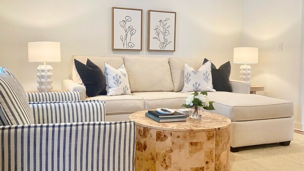 Ein modernes Wohnzimmer mit einer Couch, einem gestreiften Sessel und einem runden Couchtisch. Zwei Wandleuchten und ein gerahmtes Blumenbild hängen über der Couch.