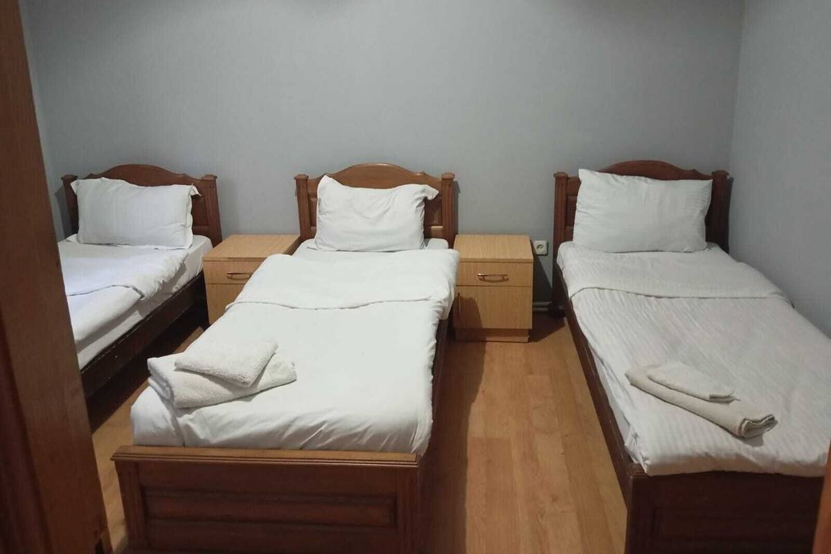 A room with three single beds, each with wooden headboards and white bedding. There are wooden nightstands with drawers between the beds.
