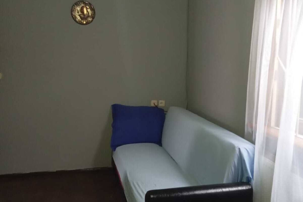 A small, sparsely furnished room with a single bed, a blue pillow, a wooden ceiling, and a window with sheer curtains.