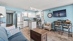 A modern kitchen with white cabinets, a black countertop, and stainless steel appliances. There is a dining area with a wooden table and two chairs. A sofa with patterned cushions is in the living room, and a TV is mounted on the wall. - Ocean Forest Plaza 606 -1BR/1BA- Pool (Myrtle Beach)