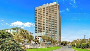 A tall, modern hotel with multiple floors and balconies, surrounded by greenery and a clear blue sky. - Ocean Forest Plaza 606 -1BR/1BA- Pool (Myrtle Beach)