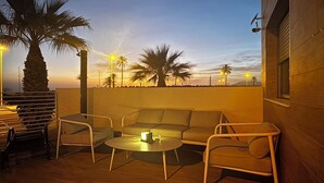 An outdoor seating area with a sofa, chairs, and a table, overlooking a sunset with palm trees and streetlights. - Calanegra Apartment with Private Pool - Ground Floor - Puerto de Mazarrón (Mazarrón)