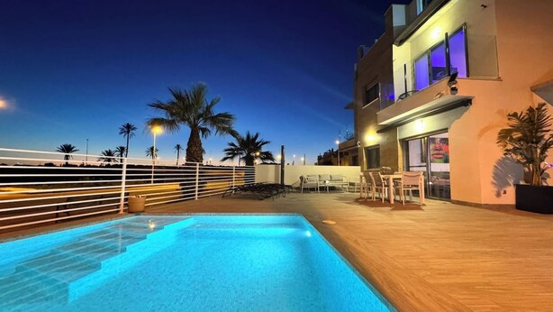A rooftop pool area with a wooden deck, lounge chairs, and a view of palm trees and buildings under a twilight sky. - Calanegra Apartment with Private Pool - Ground Floor - Puerto de Mazarrón (Mazarrón)