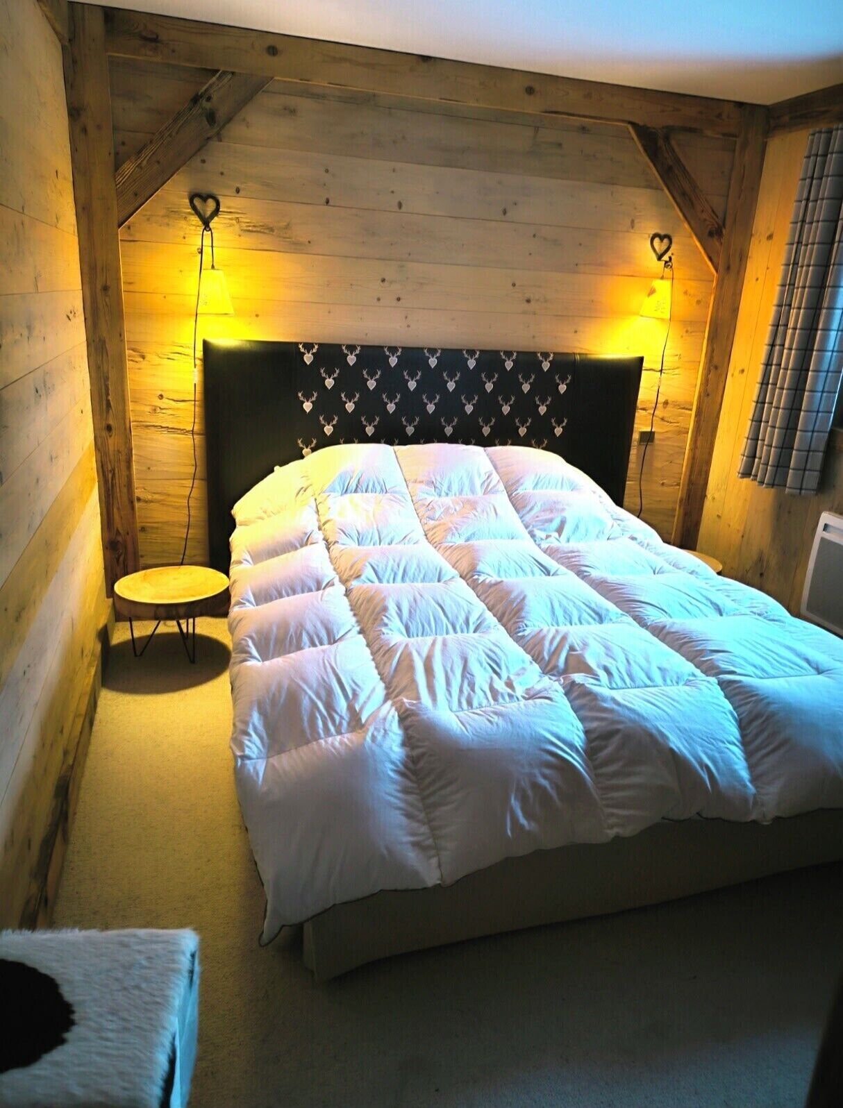 Un dormitorio acogedor con techo de madera, una cama con cabecera estampada con corazones y dos lámparas de noche.