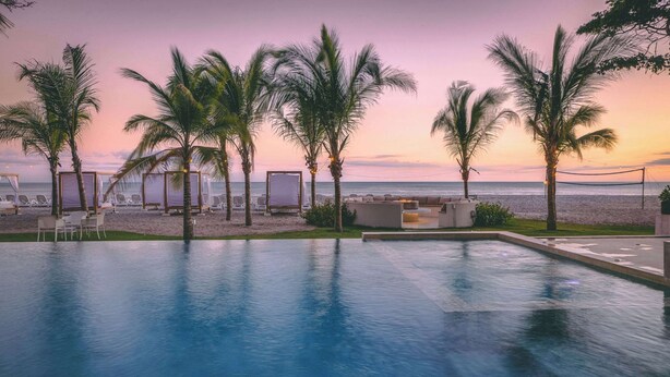 Un espace aménagé au bord de la piscine, avec des chaises longues et des palmiers, offrant une vue sur l’océan au coucher du soleil.