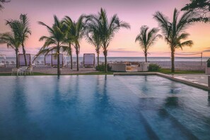 A poolside area with lounge chairs and palm trees, overlooking the ocean at sunset.