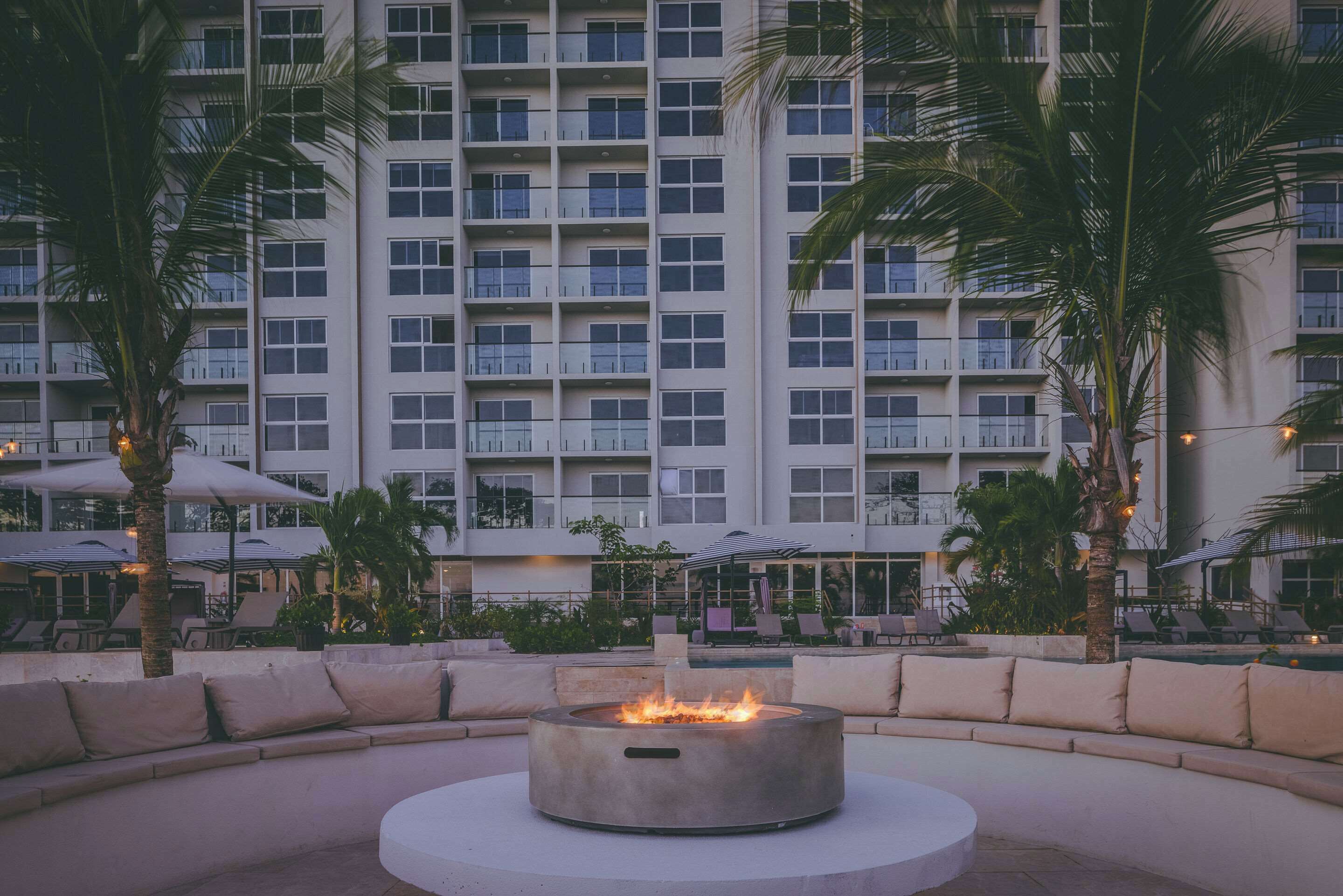 Un espacio de estar al aire libre circular con una chimenea en el centro, rodeado de sillones de descanso y palmeras, con un edificio de hotel al fondo.