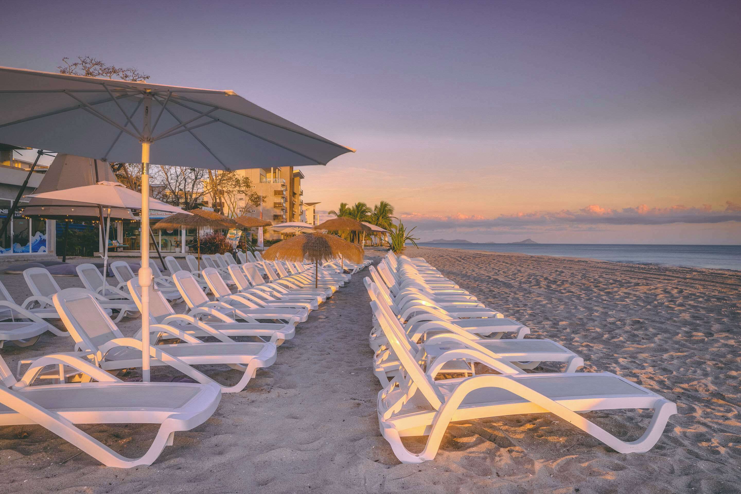 Un hôtel situé en bord de mer, avec des rangées de chaises longues et de parasols, une vue dégagée sur l’océan et un bâtiment doté de balcons.
