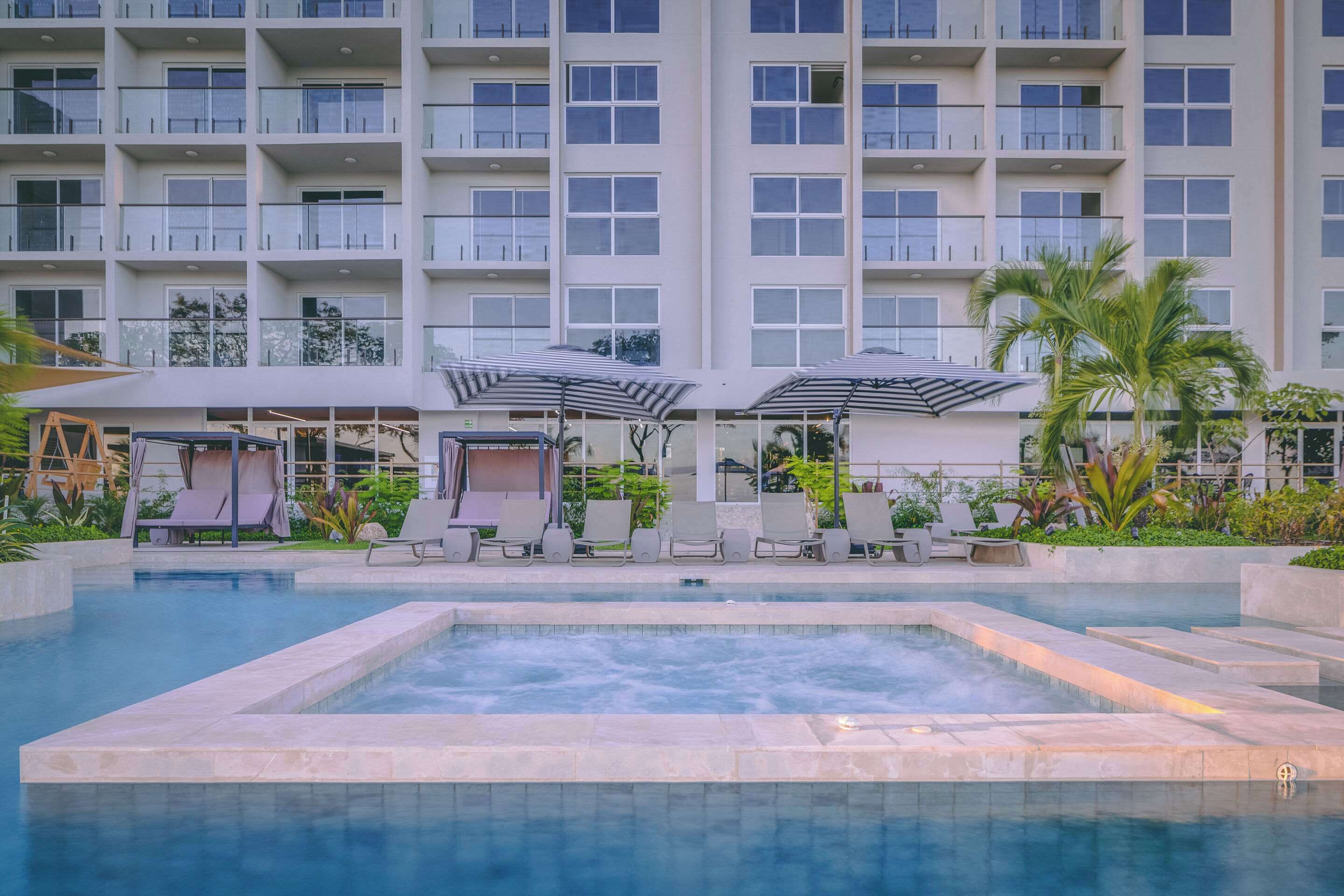 A hotel pool area with lounge chairs and umbrellas, surrounded by a multi-story building with balconies.