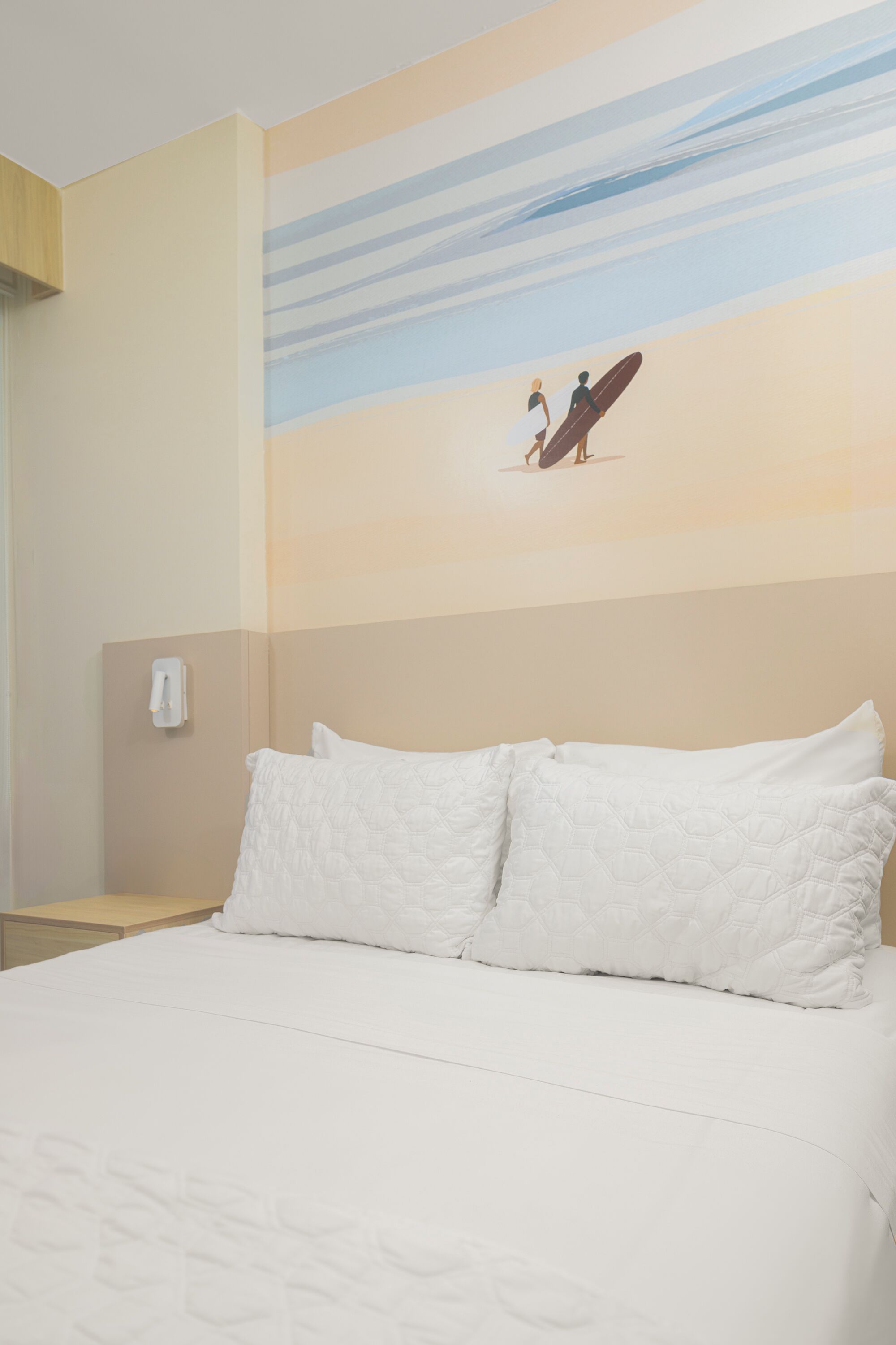 A hotel room with a bed, a nightstand, and a wall painting of two people on a beach.