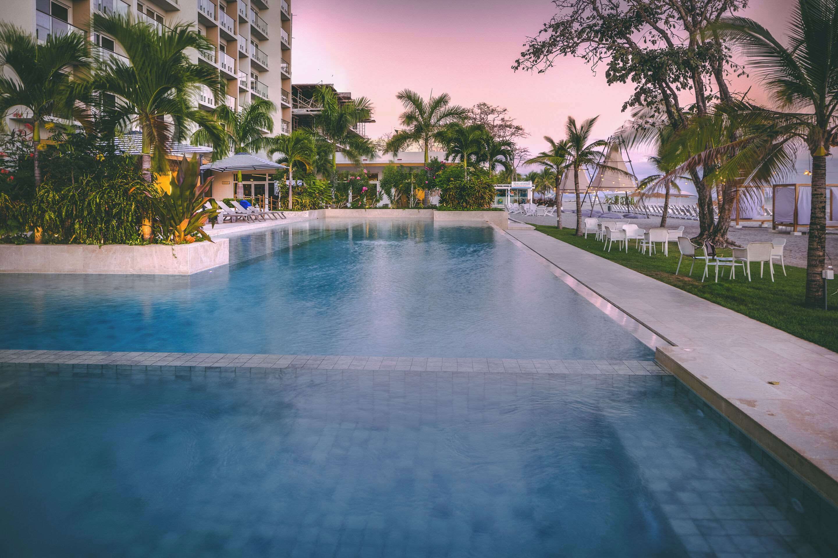 Una piscina con sillas de descanso y palmeras, rodeada por un edificio con balcones.