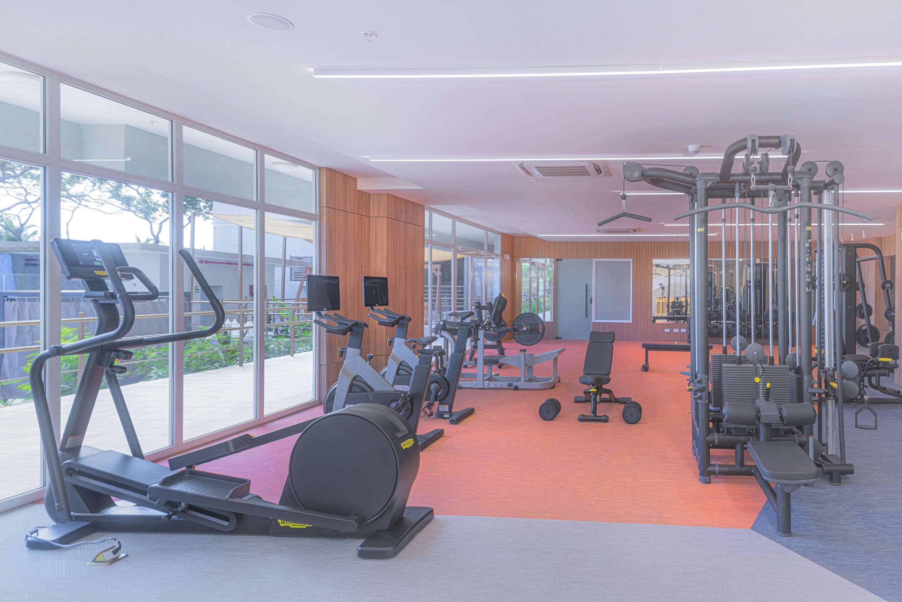 Un gimnasio con máquinas de cardio, equipo de entrenamiento con pesas y amplias ventanas con vistas al exterior.