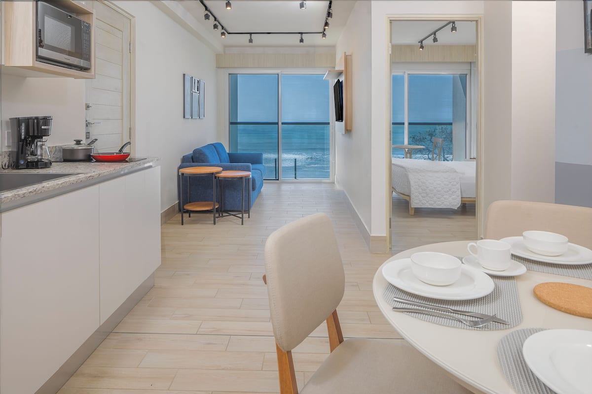 A modern kitchen with a microwave, coffee maker, and red pan. There is a dining area with a table set for two, a blue sofa, and a view of the ocean through large windows.