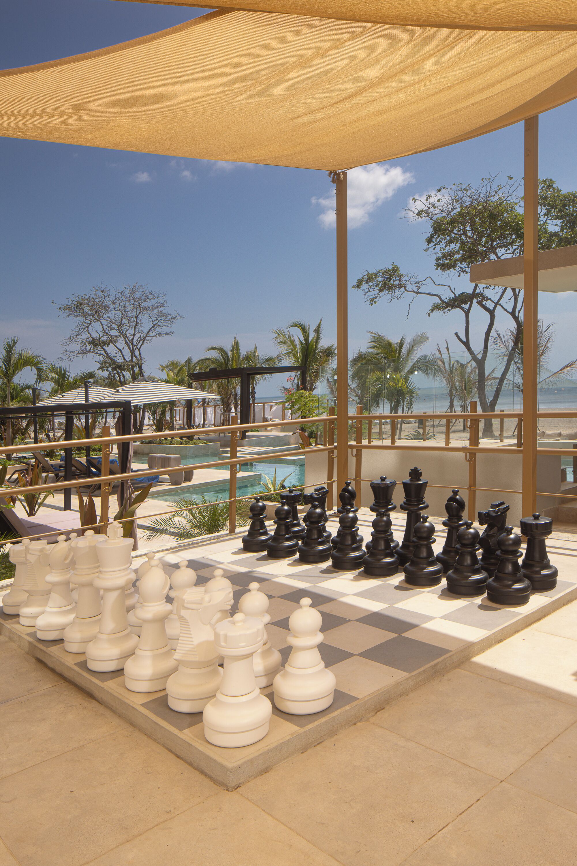A checkered chessboard with chess pieces set up outdoors, a pool, and a beach view in the background.