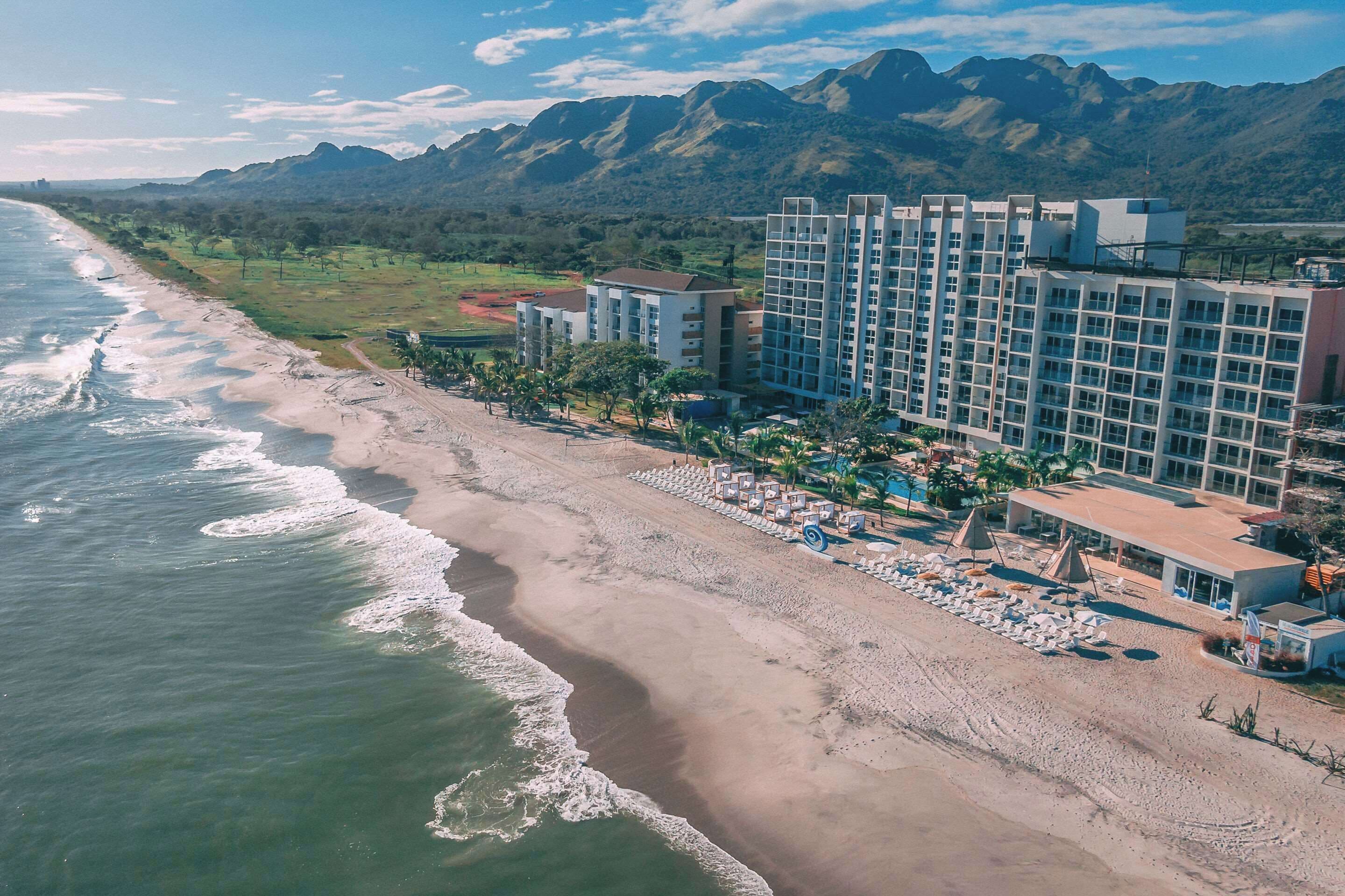 A beachfront hotel with multiple buildings, a sandy beach, and mountains in the background.