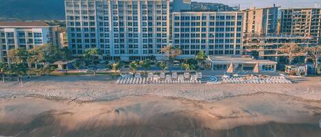 Un hôtel en bord de mer, avec plusieurs étages, une plage de sable devant et une montagne en arrière-plan.