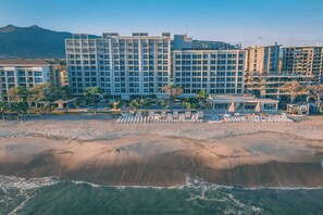 A beachfront hotel with multiple floors, a sandy beach in front, and a mountain in the background.