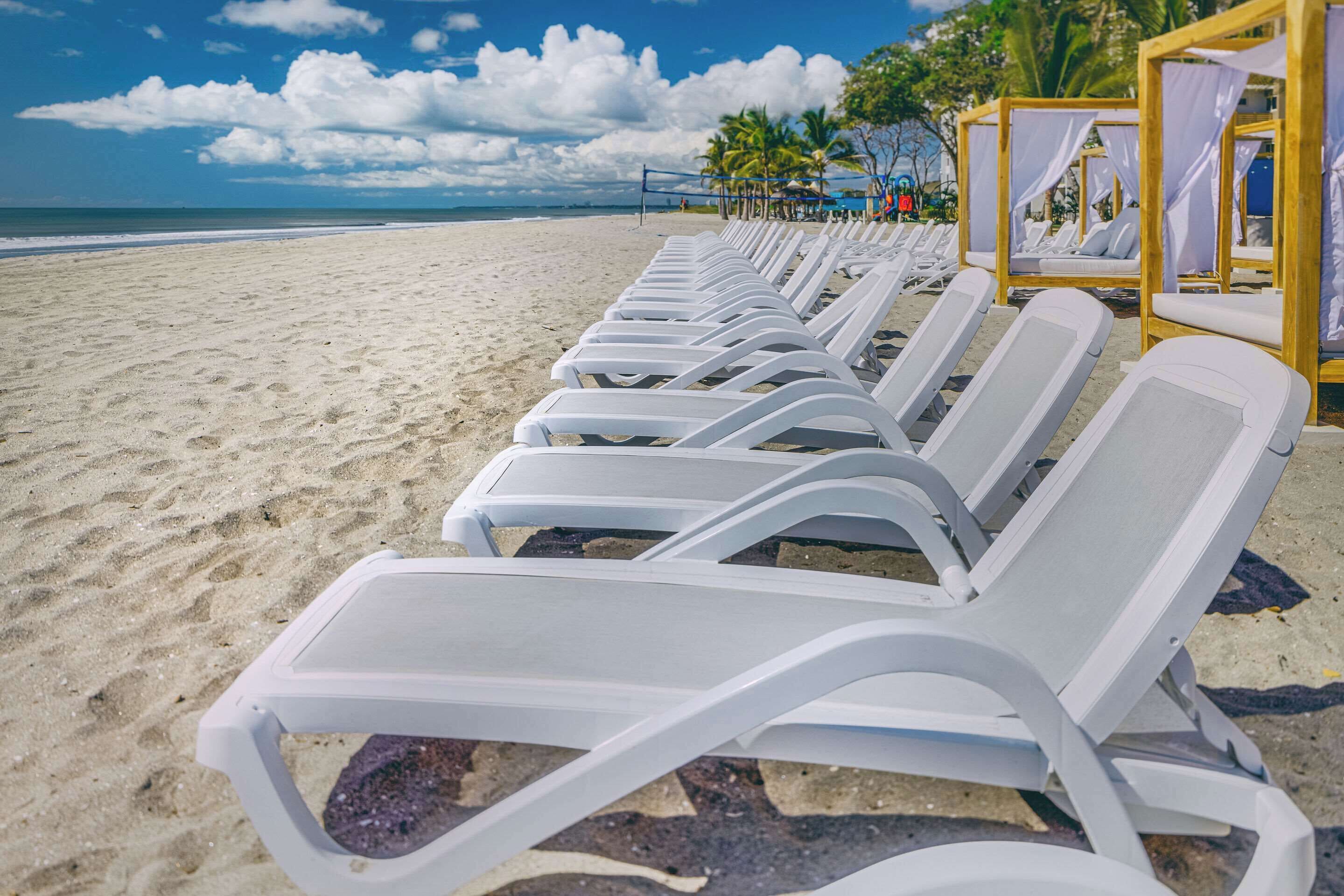 White lounge chairs lined up on a sandy beach with a clear sky and ocean in the background.