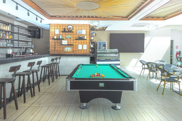 A pool table in a bar area with stools and a bar counter with bottles and glasses.
