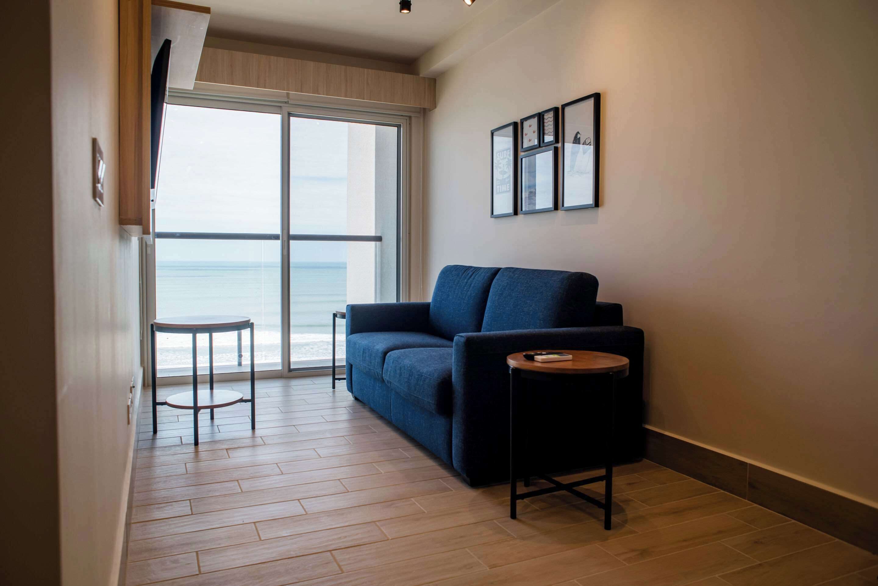 A modern living room with a sofa, a small round table, and a large window offering a view of the sea.