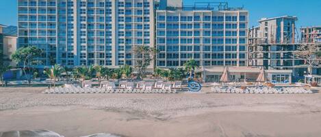A beachfront hotel with multiple high-rise buildings, a sandy beach, and a pool area with lounge chairs.