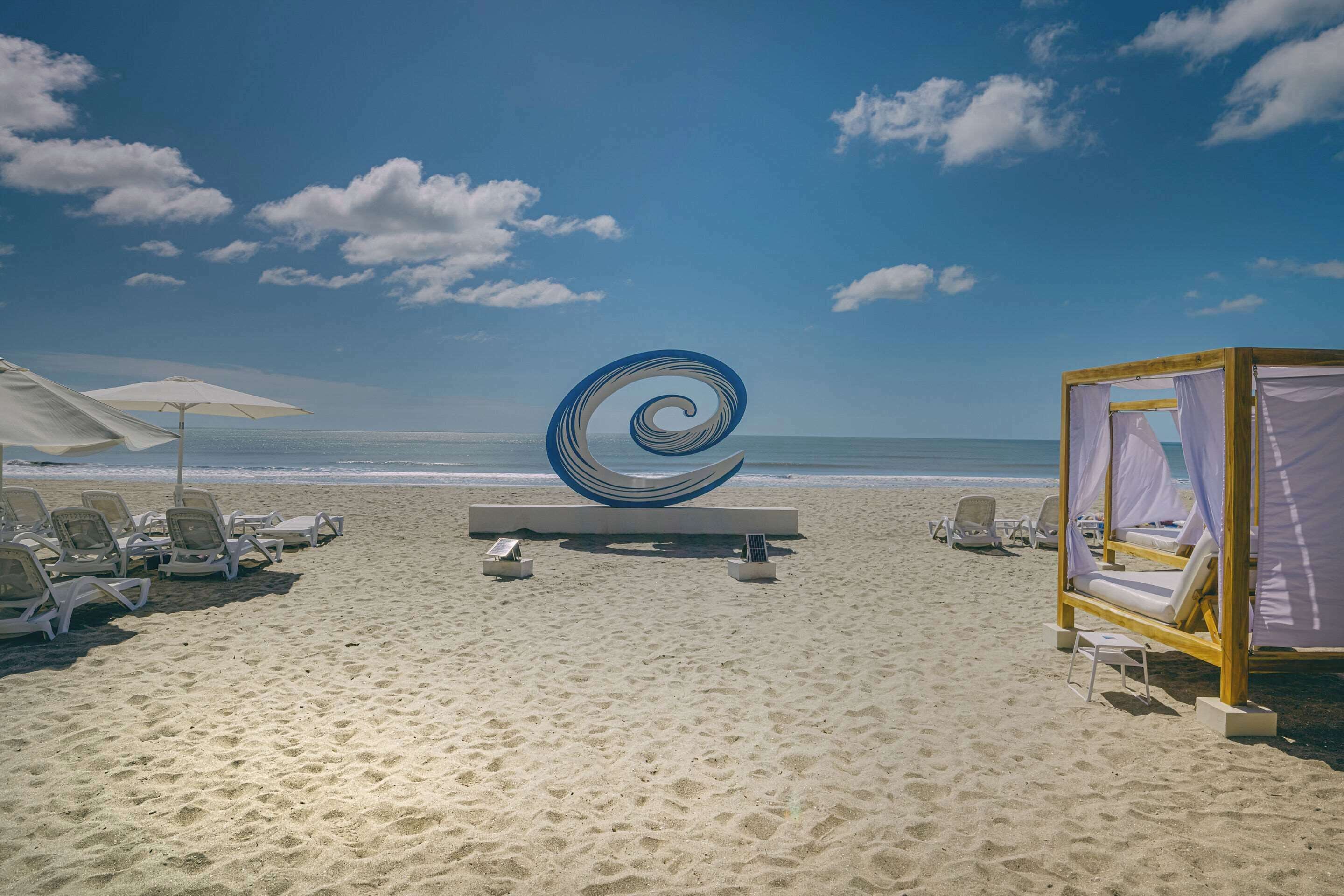 Una playa con tumbonas, una gran escultura espiral y una cabaña de madera con cortinas blancas.