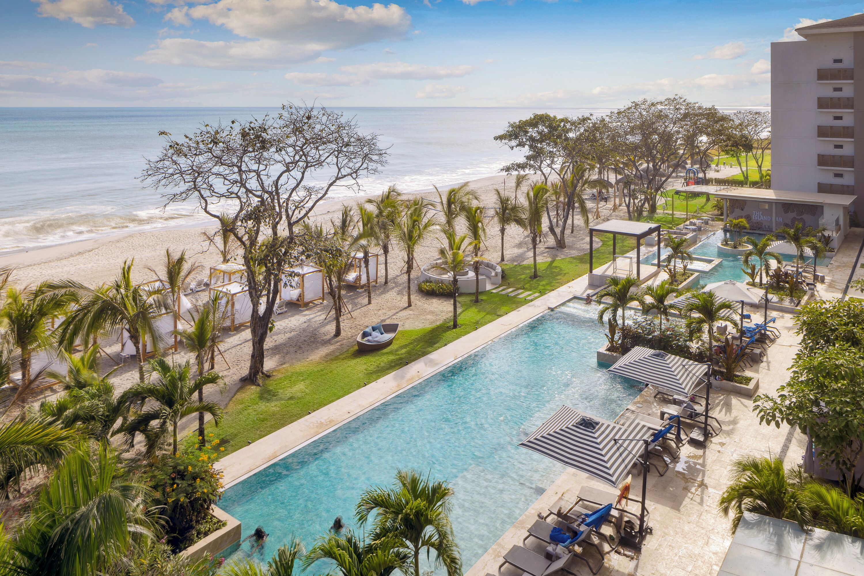 A resort pool with a view of the beach, surrounded by palm trees and lounge chairs.