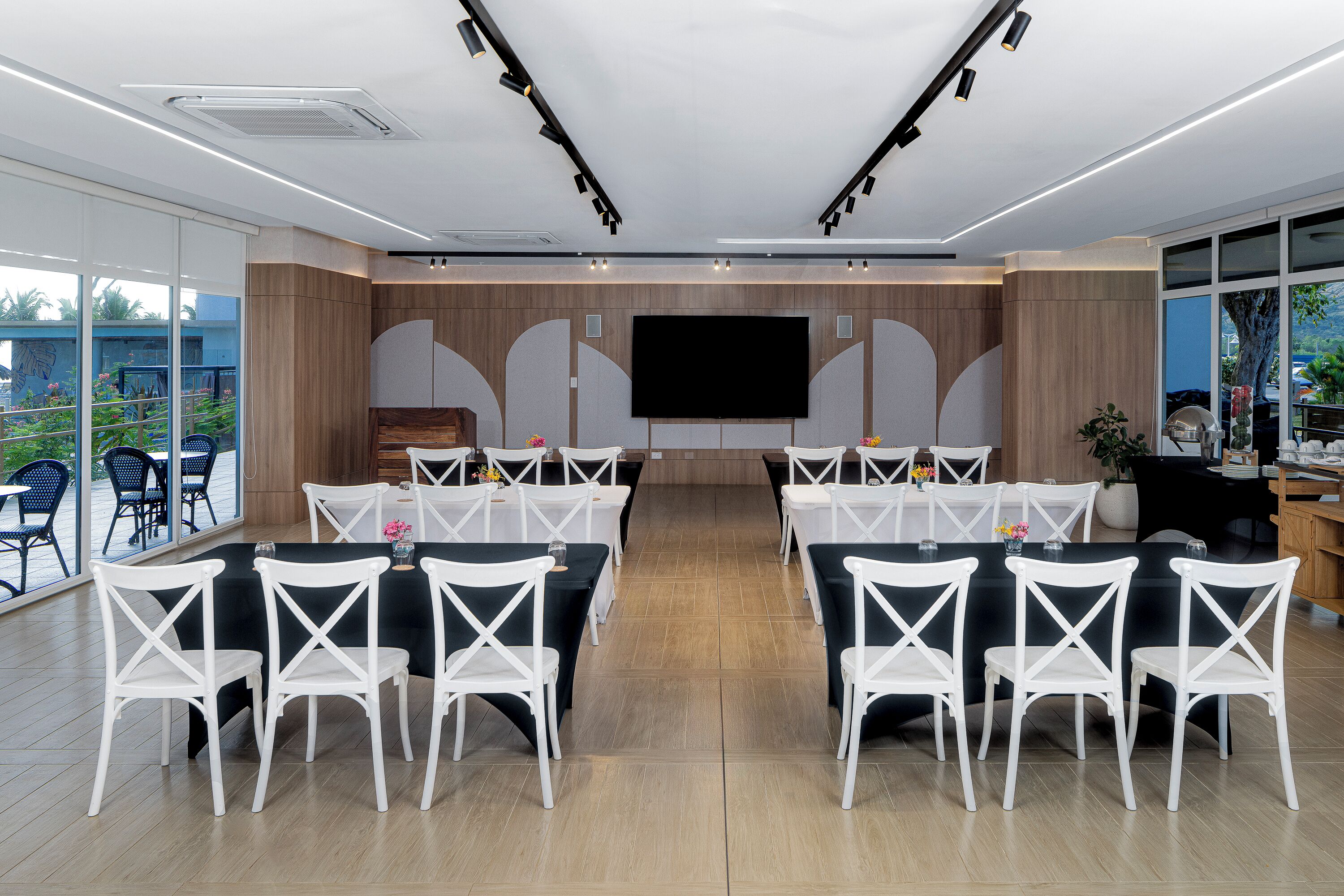 A modern conference room with white chairs and tables, a large flat-screen TV, and a balcony with outdoor seating.