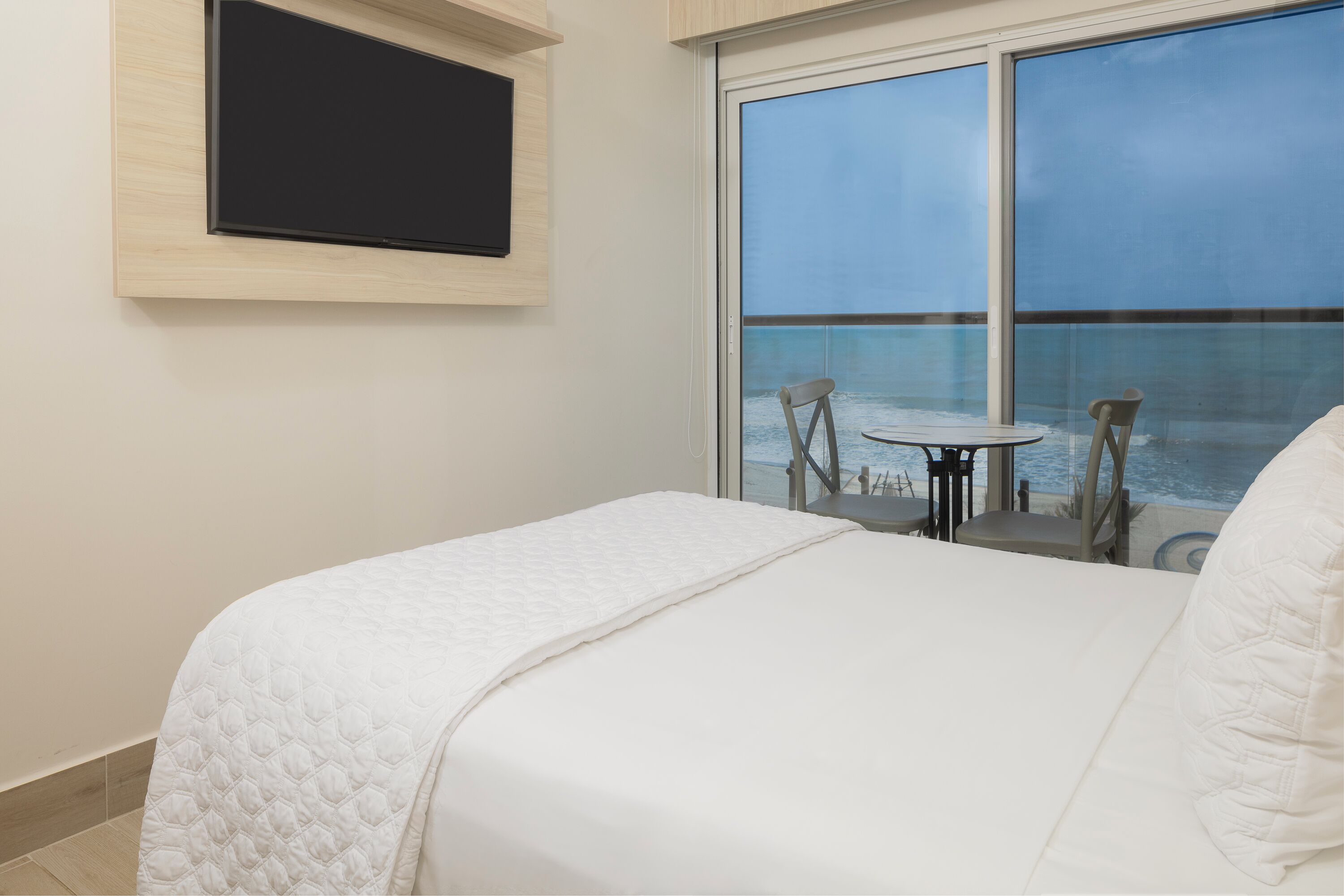 A hotel room with a bed, a TV mounted on the wall, and a balcony with a view of the sea.
