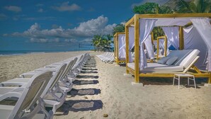 Une plage bordée de rangées de chaises longues et de cabanes en bois aux rideaux blancs.