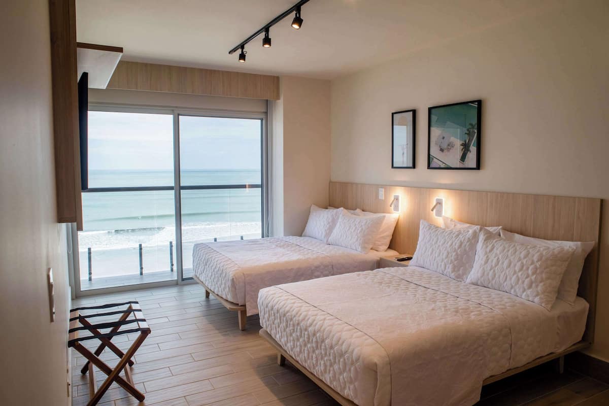 A hotel room with two beds, a large window with a view of the sea, and a small table.