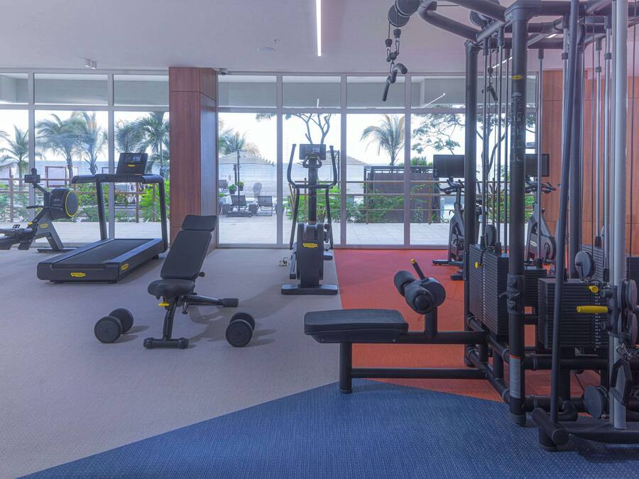 A gym with various exercise equipment, large windows, and a view of palm trees and a beach.