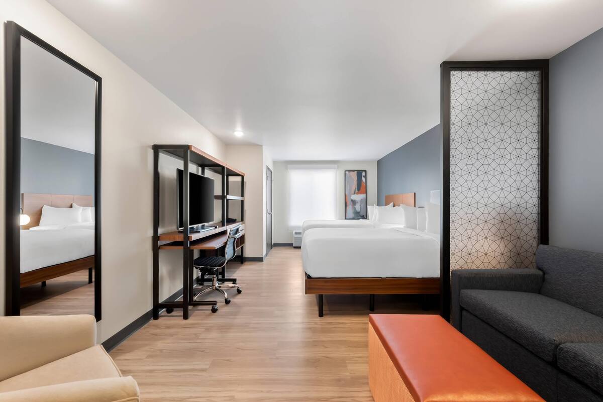 Quarto de hotel com cama, escrivaninha com computador, cadeira, sofá e um espelho grande.