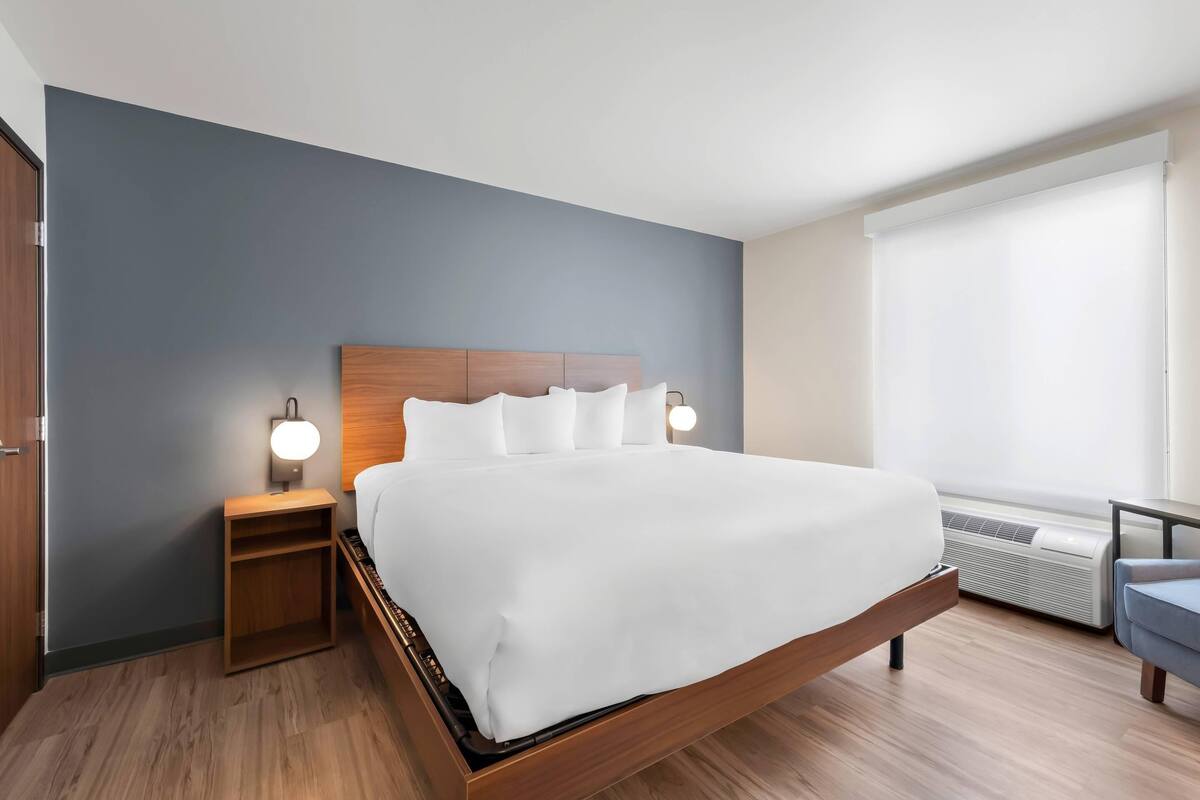 Quarto de hotel moderno com uma cama grande, mesas de cabeceira e uma janela com persianas.