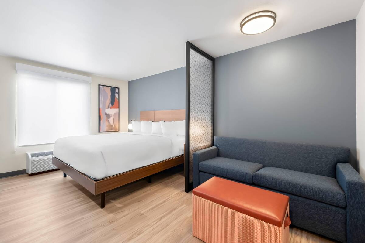 Quarto de hotel moderno com uma cama, um sofá cinzento e um banquinho laranja pequeno.