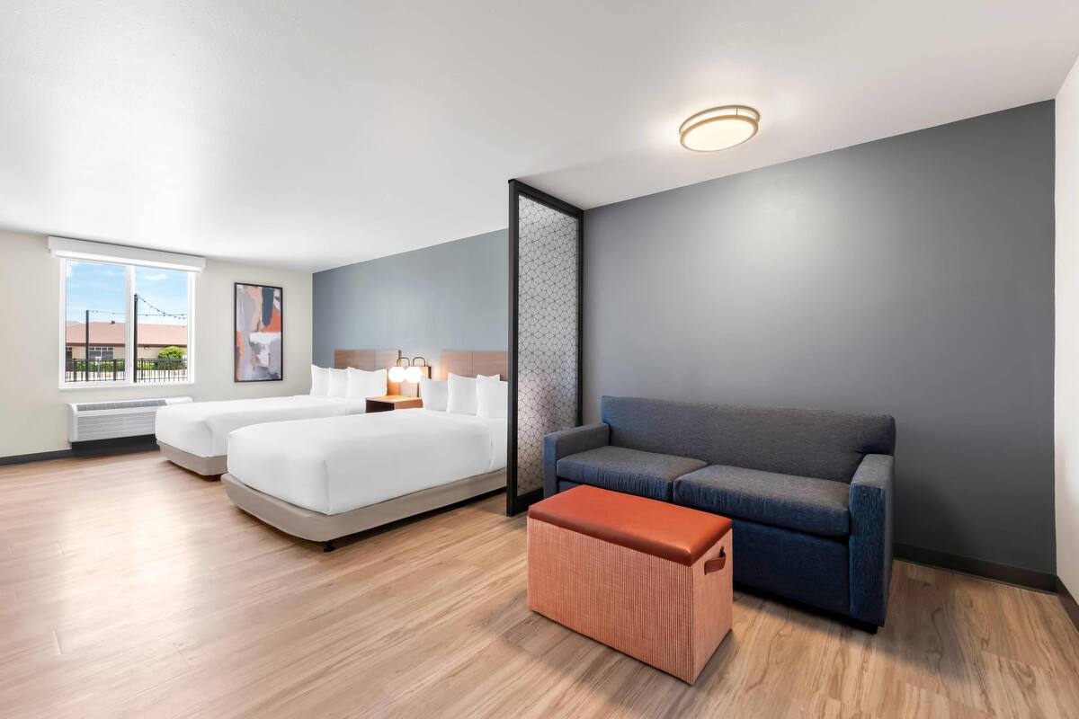 Quarto de hotel moderno com uma cama grande, um sofá, um pequeno banquinho e uma janela com vista para edifícios.