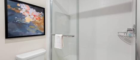 A modern bathroom with a glass shower enclosure, a white toilet, and a framed abstract painting on the wall.