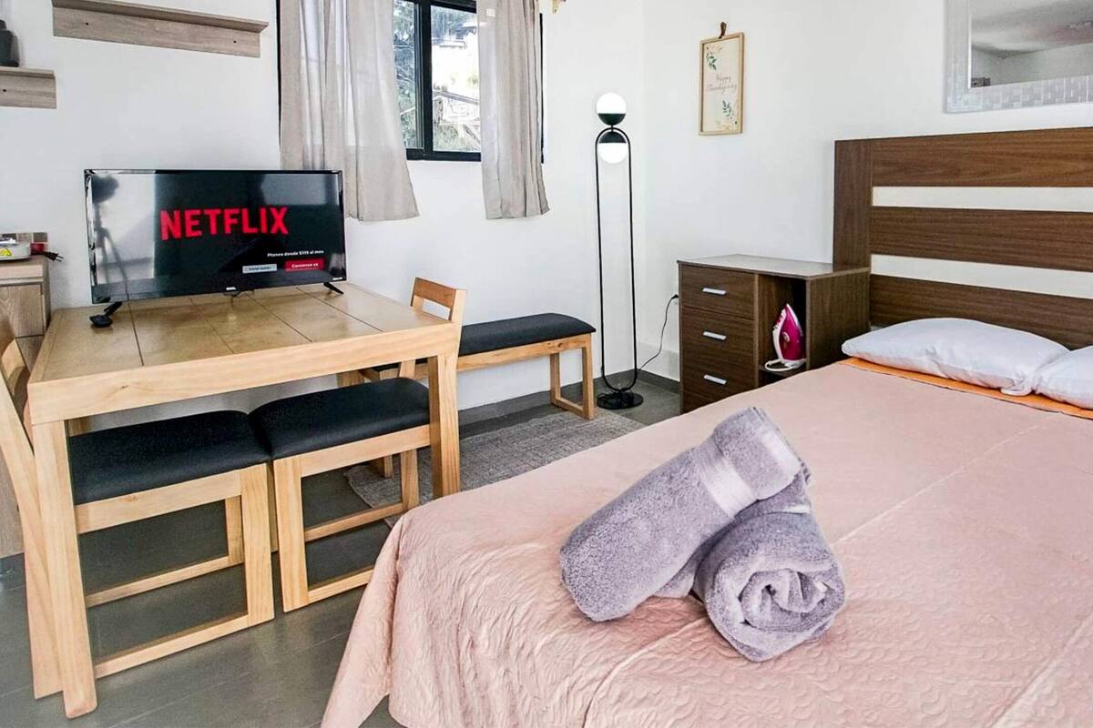 A neatly arranged bedroom with a bed, a desk with a laptop, a TV displaying 'NETFLIX', and a bedside table with a lamp.