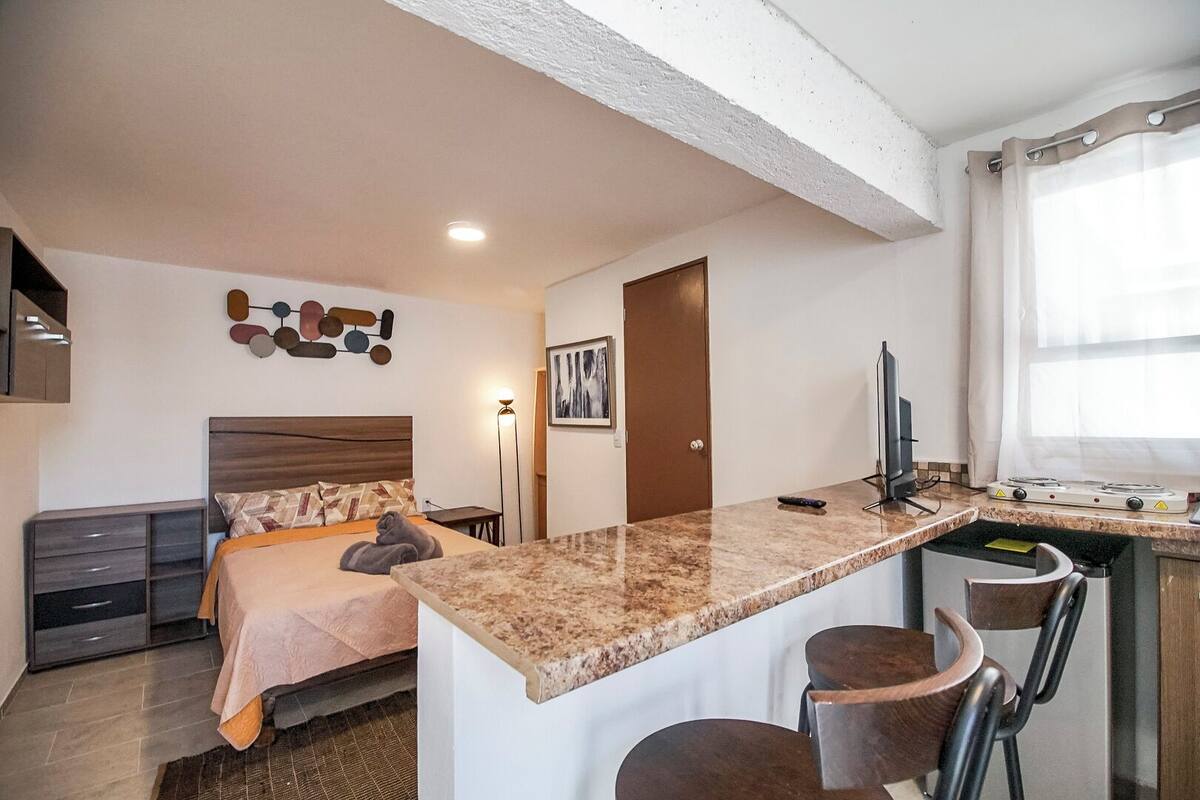 A compact hotel room with a bed, a small desk, a chair, a kitchenette, and a bathroom.